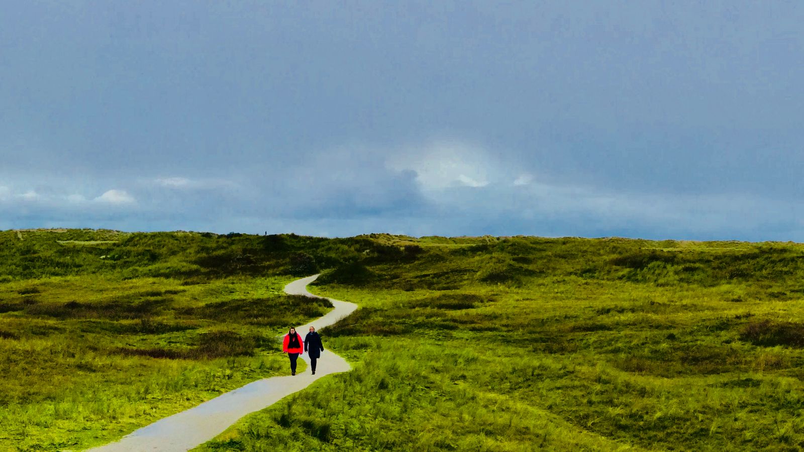 A pale gravel footpath winding diagonally across green dune grassland under a wide, moody blue sky, with two people walking along it in the distance