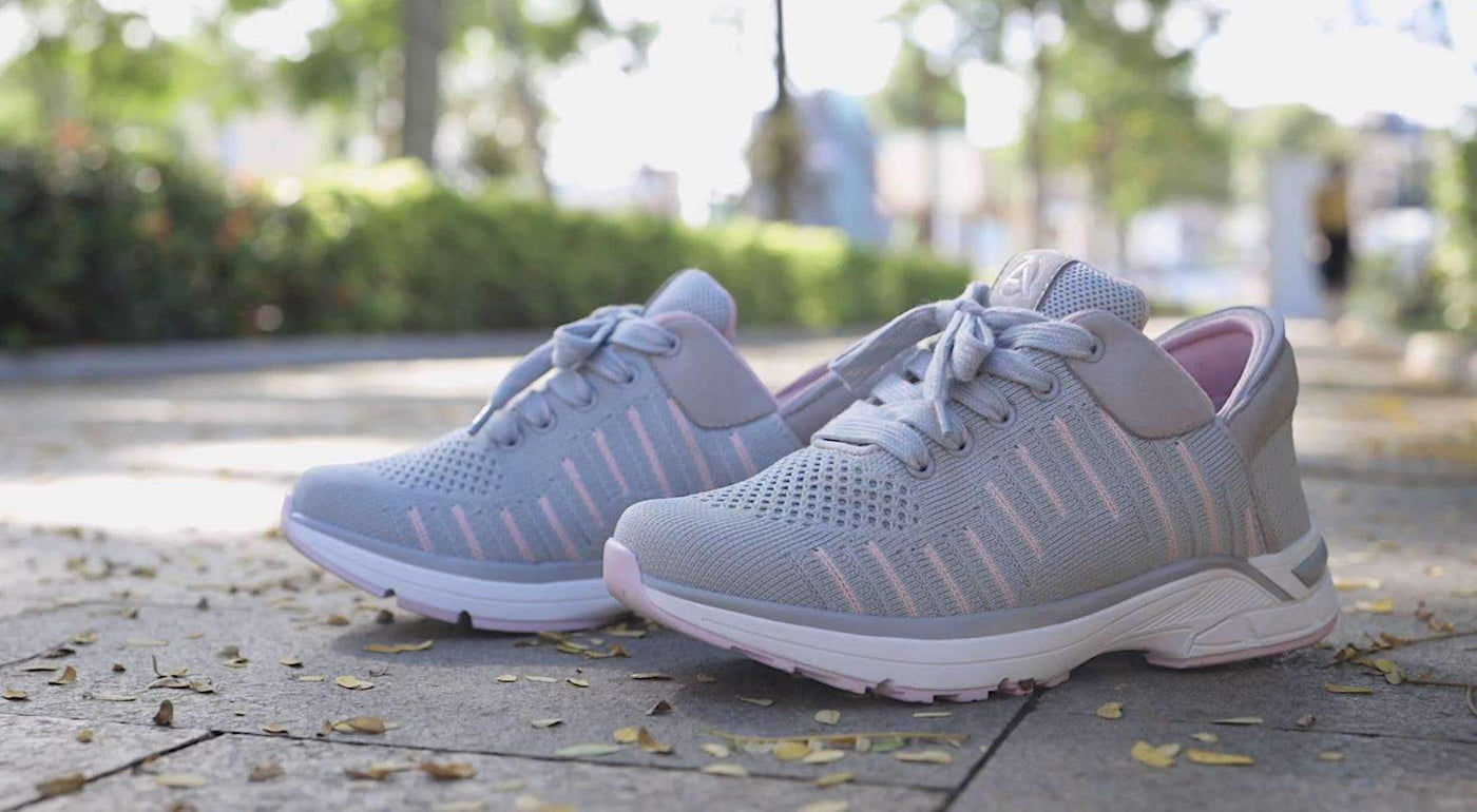 A pair of Zeba Rose Gray knit sneakers in grey with pink stitching, standing on a paved path with a blurred hedge behind